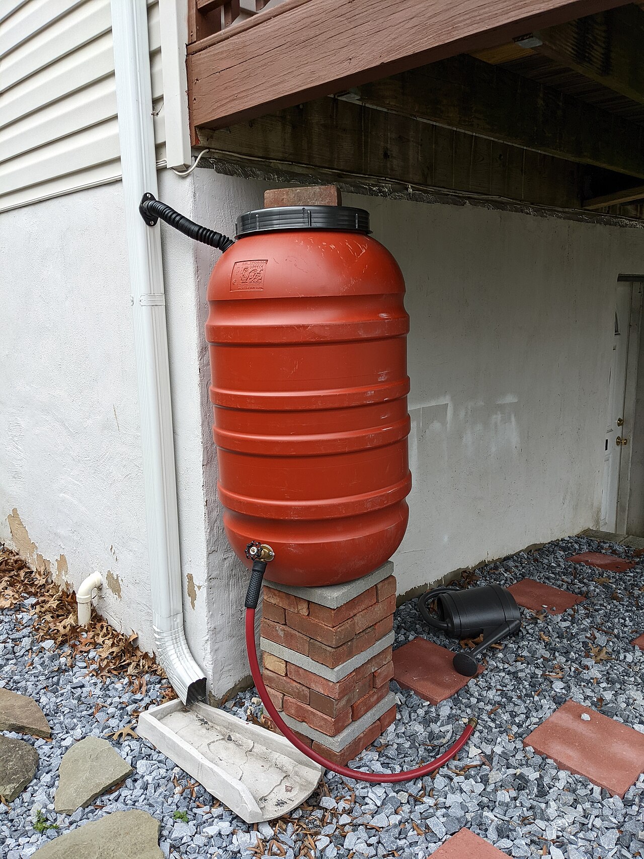 Red rain barrel connected to a roof downspout on a brick base